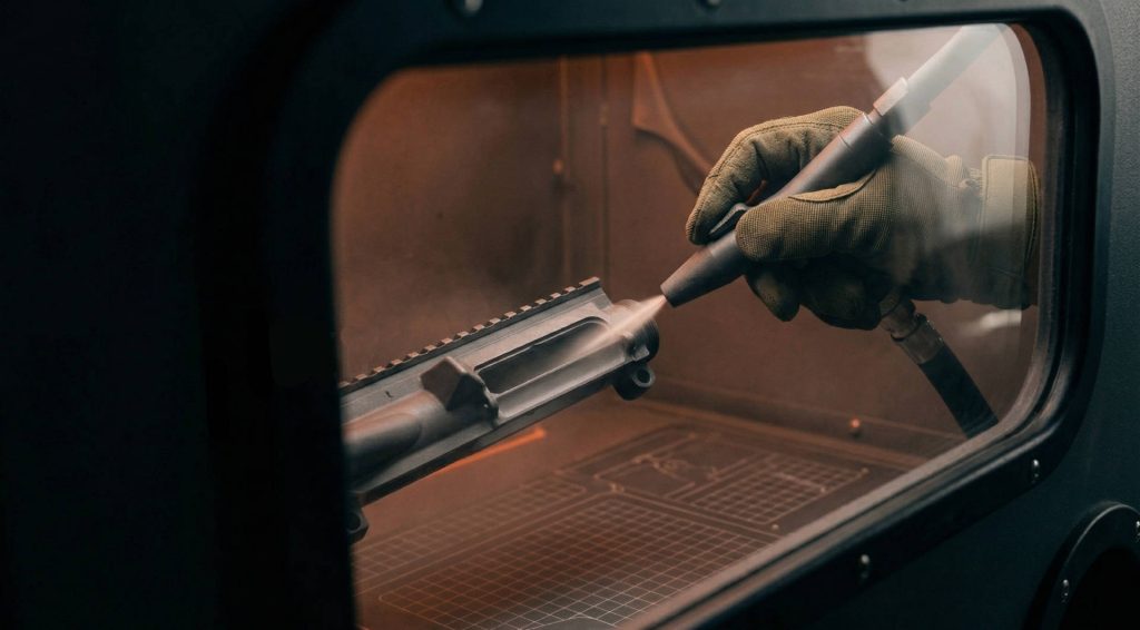 An industrial macro shot looking through the window of a media blasting cabinet. A gloved hand directs a precise stream of fine media at an AR-15 upper receiver, removing old finishes. Subtle orange light illuminates the cabinet interior.