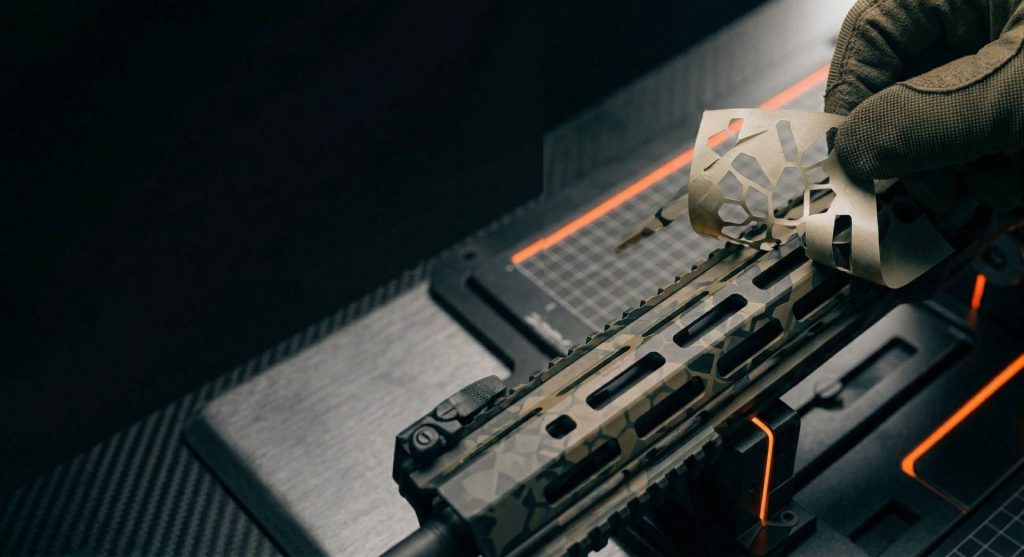 A macro close-up of a gloved gunsmith hand carefully peeling a vinyl stencil layer from an AR-15 handguard. The process reveals an intricate, layered geometric Hex pattern in multiple Cerakote colors.