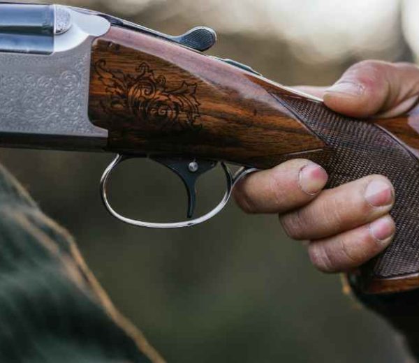 An ornate hunting shotgun is held firmly in a hand. The weathered wooden handle of the gun features etched designs.